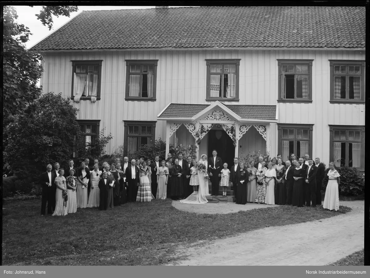 Brudepar foran bolighus med brudefølge. Brudgom er Halvor Holta. Bolighusets inngangsparti er dekorert med snekkerglede.