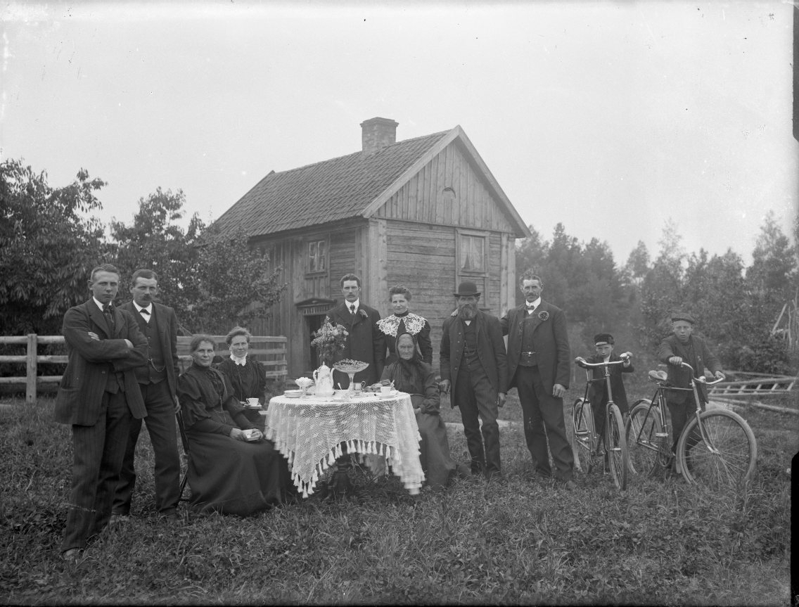 Gruppbild med sittande och stående runt ett kaffebord: ett äldre par, män, kvinnor och två barn stående vid varsin cykel. Sannolikt familjegrupp. I bakgrunden ett mindre tvåvåningshus med delvis locklistpanel.