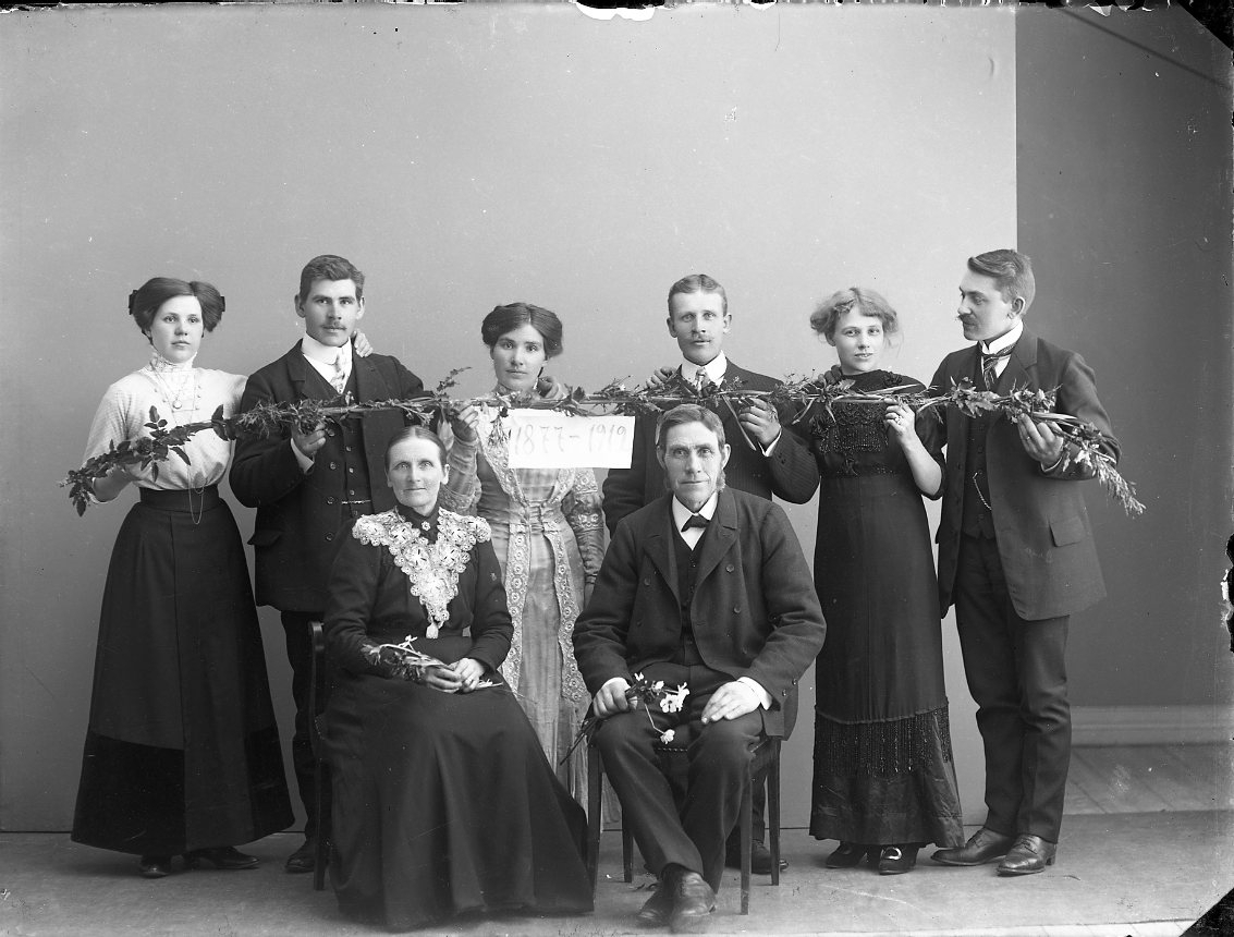Ett äldre par sitter framför sex män och kvinnor som håller en lång pinne med flätade kvistar och en lapp med texten "1877-1912". Det äldre paret firar 35-årig bröllopsdag.