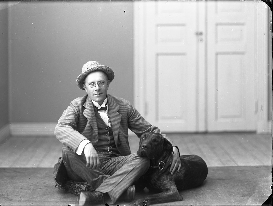 En man med glasögon sitter på golvet i Calla Sundbecks ateljé tillsammans med en hund.