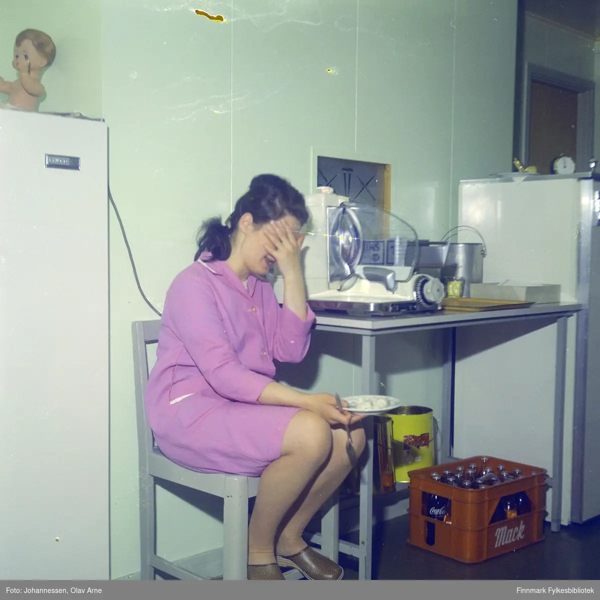 En ukjent kvinne spiser men hun sitter på en stol i et kjøkken, antagelig på Skansen i Båtsfjord

En kasse kan bli sett til høyresom inneholder coca-cola flasker

Foto trolig tatt på 1960-tallet