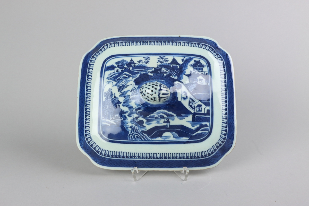 Square form with chamfered corners and slightly domed shape, on top a knob in form of a fruit. On the lid landscape scenes of pagodas, buildings,  bridges, figures with parasolls,  gardens and waters. The edge of the lid is decorated with a wide dark blue border with a criss cross pattern. All decorations in blue underglaze. The inside of the lid without decoration.