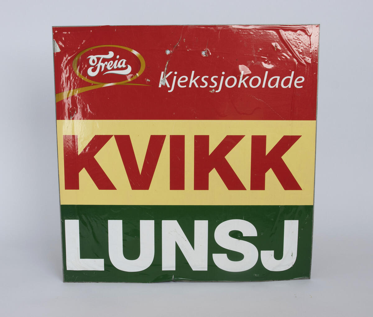 Firkantet reklameskilt for kjekssjokoladen Kvikk Lunsj med en tredeling av farger i rødt, gult og grønt, med hvit skrift. 