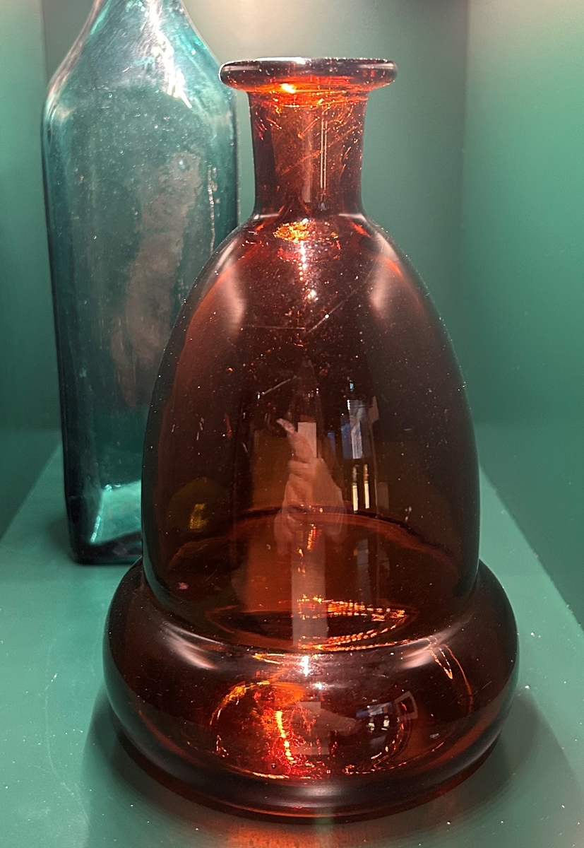 Flaska av brunt glas med rund, välvd botten. Konisk, indragen hals, utåtvikt mynningskant.