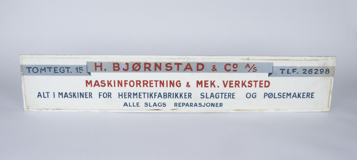 Reklameskilt for maskinforretning og mekaniskverksted H. Bjørnstad & Co A/S. Skiltet er av glass og har en hvit bakgrunn med et grått banner der bedriftens adresse, navn og telefonnummer er skrevet. Under er teksten i rødt og blått. 