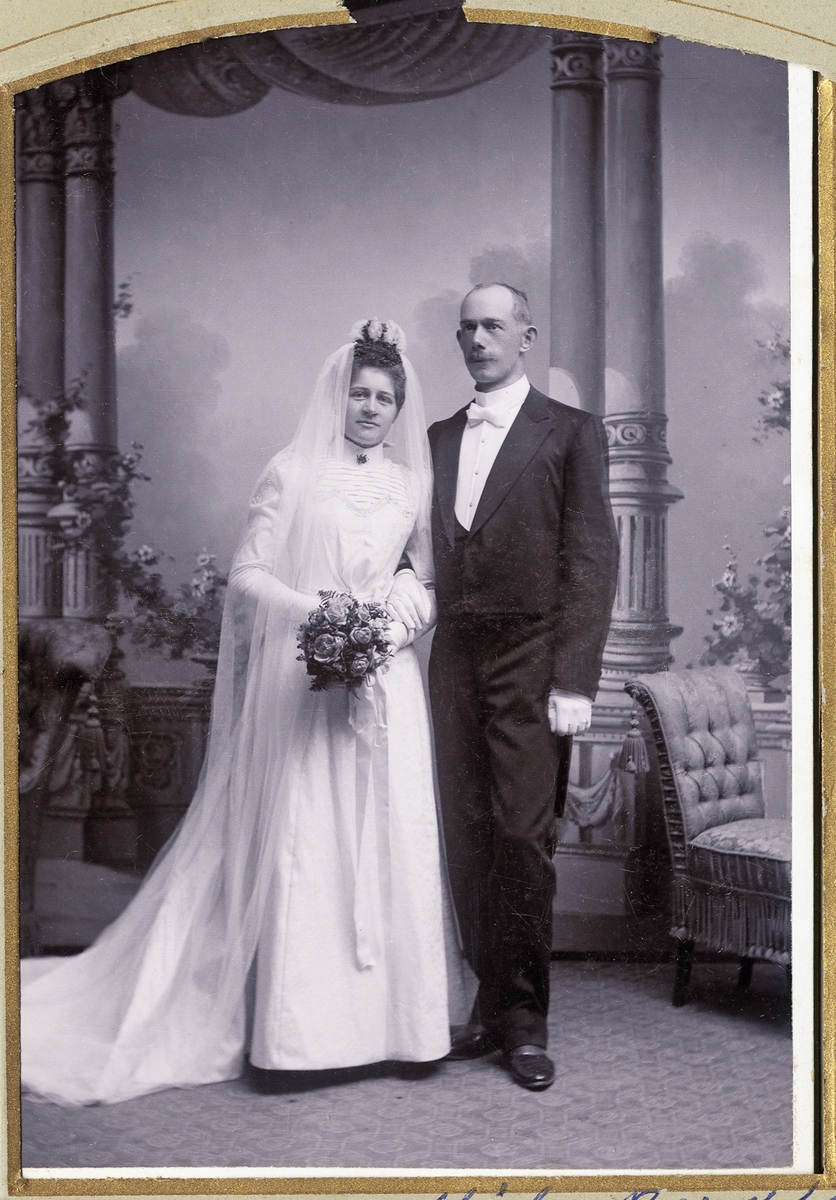 Ett brudpar. Hon i vit brudklänning med slöja och myrtenkrona, han i frack, hos fotografen, 16/7 1899.
Helfigur. Ateljéfoto.