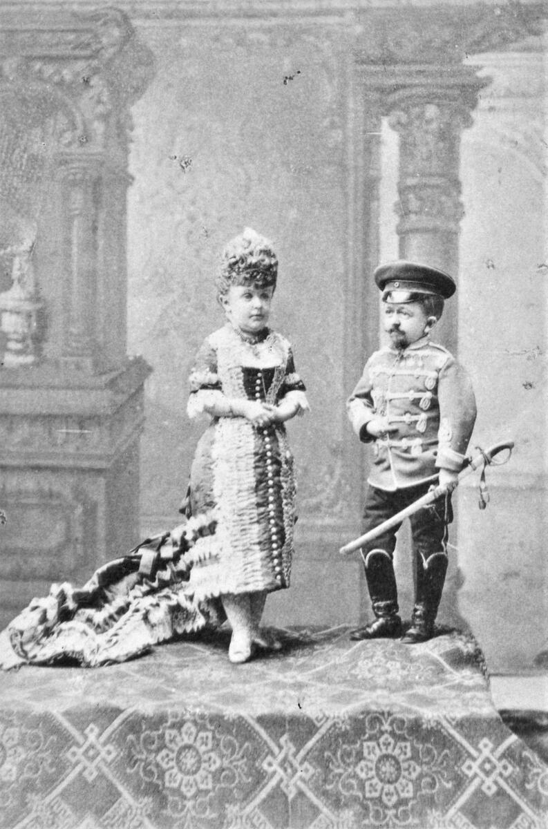 Från August Bondesons vänkrets. Ett kortvuxet par där mannen är klädd i uniform med något slags svärd och kvinnan bär en klänning med släp. Fotografiet är taget i Alexandria, Egypten.