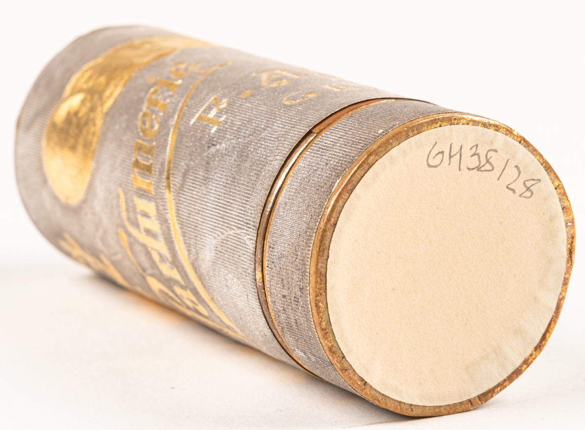 Pappersask, cylinderformad, innehållande svarvad parfymflaska i trä. Asken silverfärgad med guldfärgat tryck, invändigt röd.