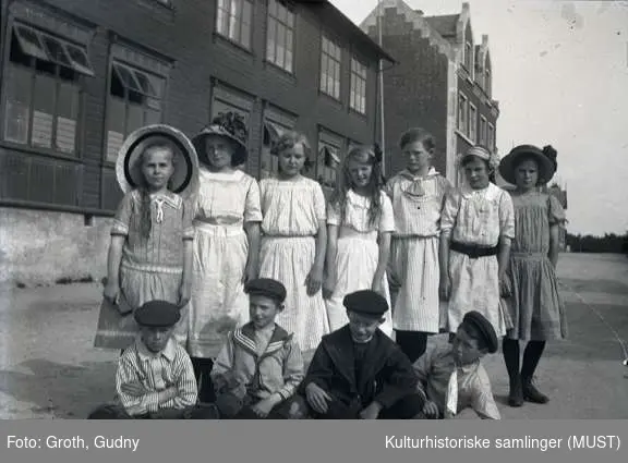 Gruppeportrett utendørs. Syv jenter og fire gutter i finklær foran Platou skole i Birkelandsgaten (tidligere Frk. Rings skole).