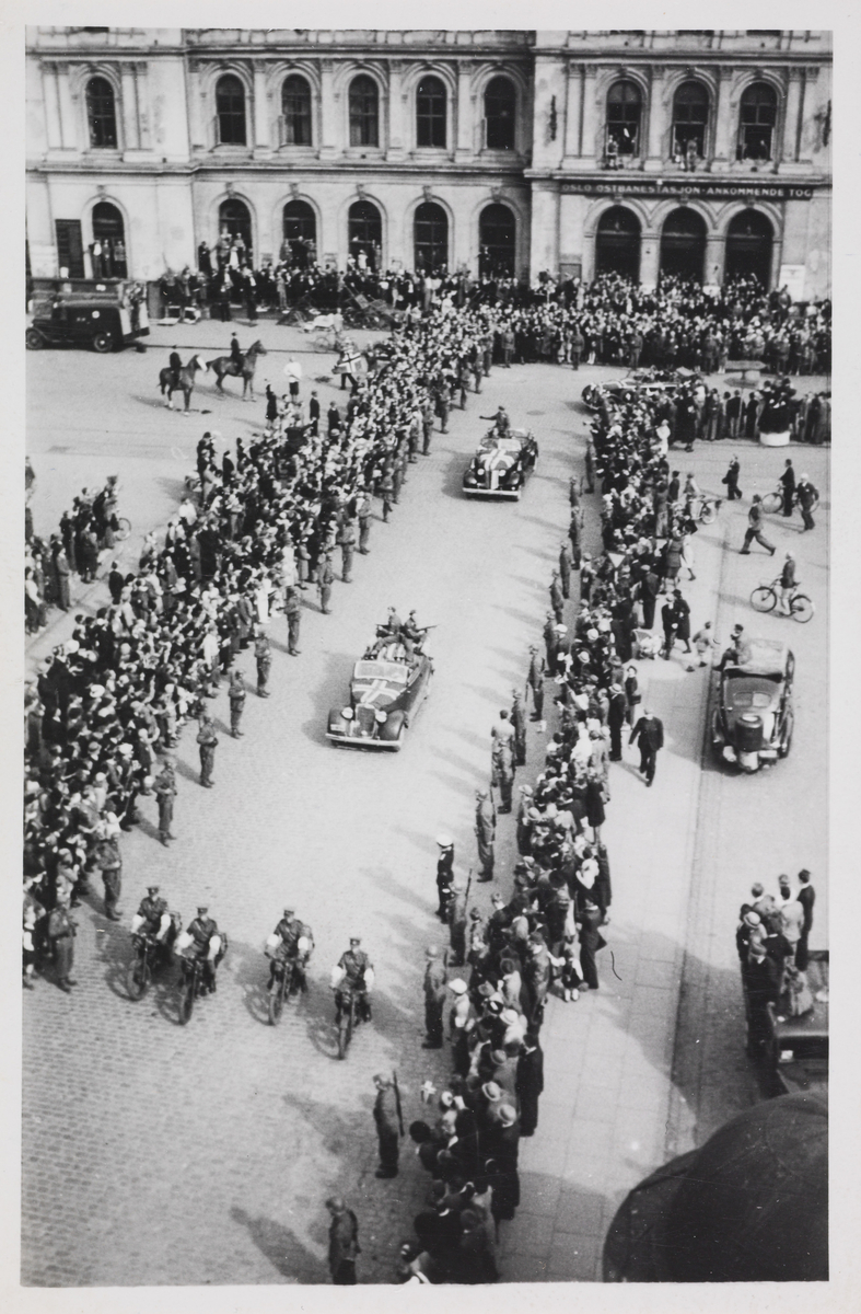 Del av bilkortesjen til Kronprins Olav, 13. mai 1945. De er på vei opp til Slottet. Bildet er tatt ved Østbanehallen. I bil nummer to bak motorsyklene, ser man Kronprinsen vinke til folkemengden. I setet foran ham sitter Max Manus som livvakt.