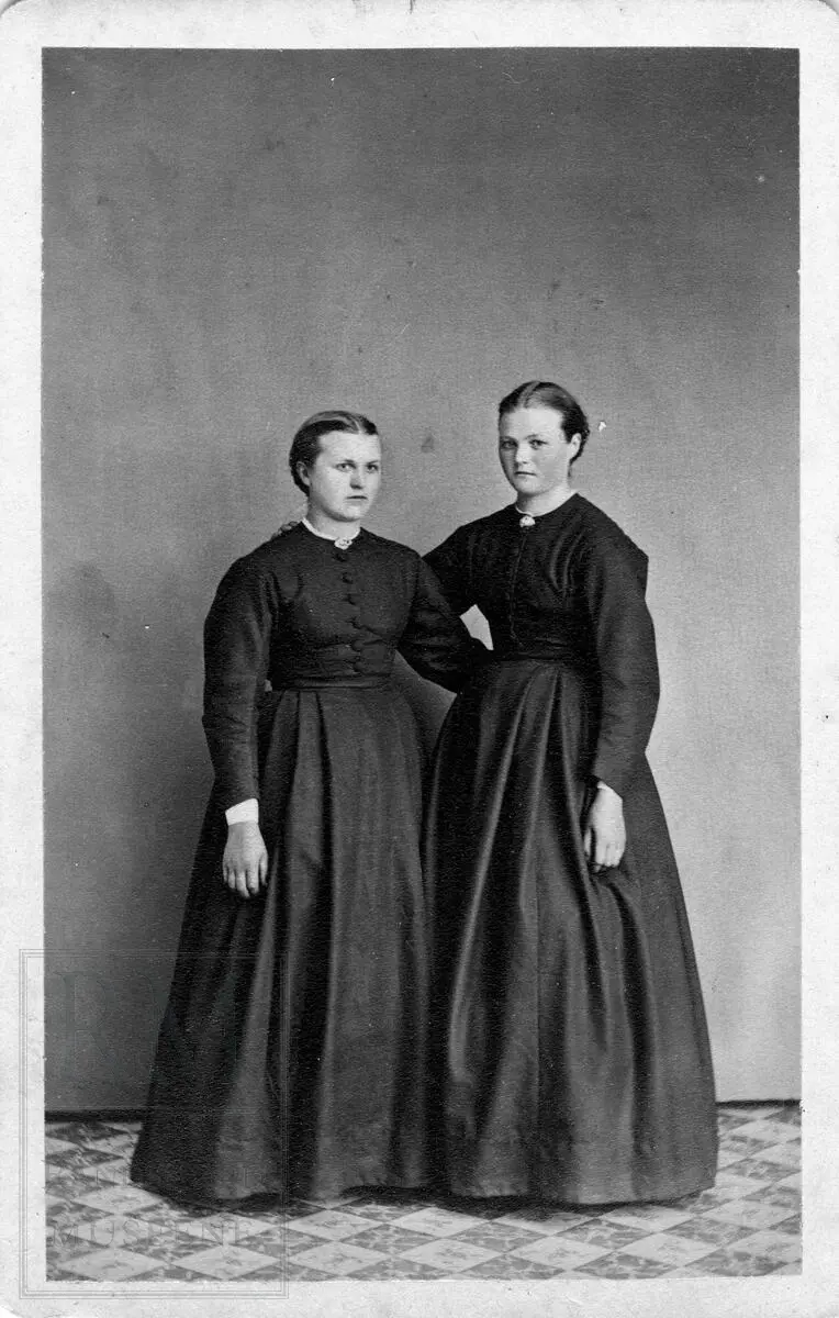 Portrett av to kvinner i helfigur. De har mørke fotside kjoler.