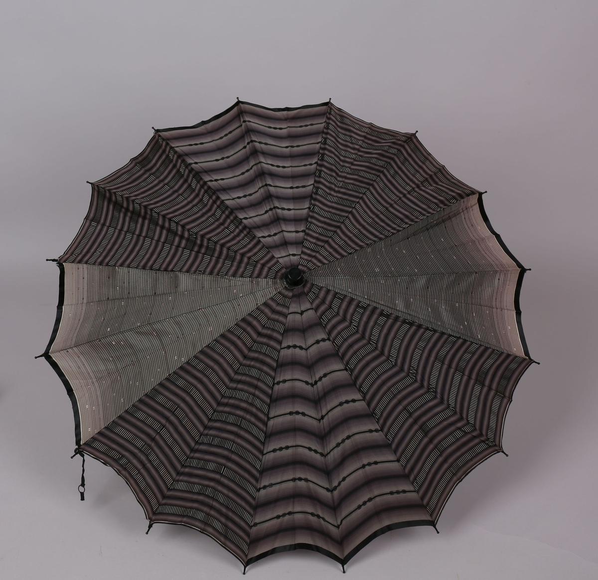 Paraply med 16 metallspilar og stong i tre. Trekt med stoff i kunstfiber, fire ulike mønstre, to og to felt i same mønster. Handtak i tre med hempe til å ta inn på handa. Hempe for lukking av paraplyen.