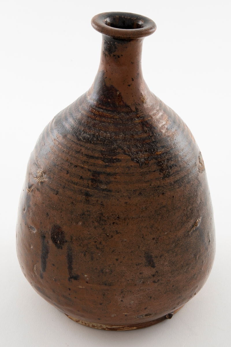 Pæreformet vinflaske med trang hals og utbrettet kant. Foten er vid med bred rand. Flasken er glasert i brun med nyanserte sjatteringer.