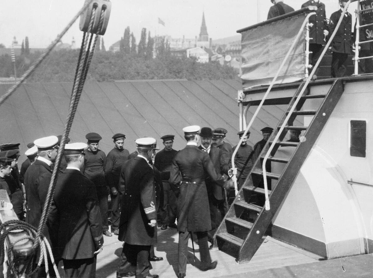 Kong Haakon mottas på dekk av gruppe menn i uniformer og gaster mens skipet ligger ved kai i Oslo. Bildet er del av en serie tatt i forbindelse med passasjerskipet "Kristianiafjords" første seilas mellom Oslo og Bergen 4.-7. juli 1913. 
