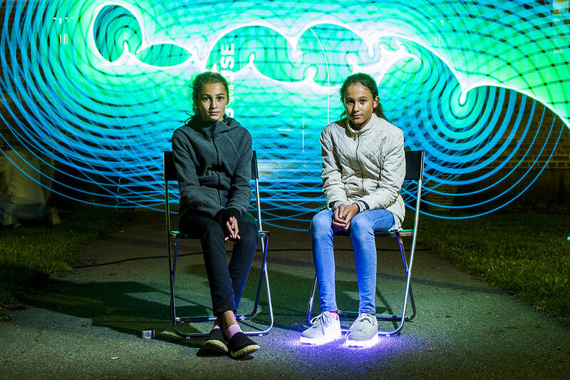 To jenter sitter på hver sin stol og ser mot fotografen. Bak dem er det et lysende mønster i grønt og blått.