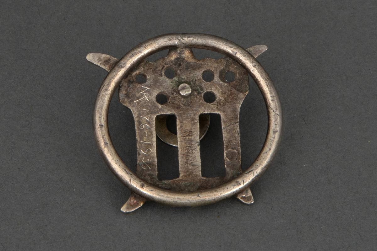 Prydmalje i forgylt sølv. Kraftig ring med en påloddet gjennombrutt, og muligens, en sterkt stilisert utforming av kronet gotisk minuskel "m". Foran en bøyle med en nedhengende flat ring.