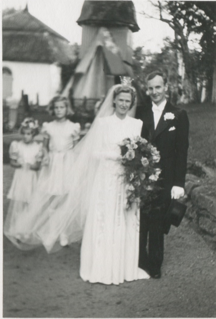 Det nygifta brudparet Ulla Hedvall, Kållered, och Harry Fredriksson (1905 - 1986), Livered, står utanför Kållereds kyrka 1942-08-15. I bakgrunden ses två brudnäbbar samt klockstapeln.