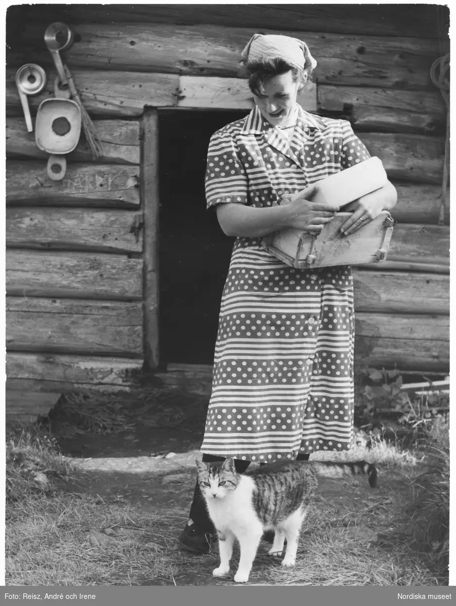 Jämtland. En kvinna som  tillverkar ost står vid en fäbodvall i Ottsjön med ost och ostkar i famnen. Hon har en katt vid sina fötter. Flera redskap för ostframställning hänger på husväggen till vänster i bild.