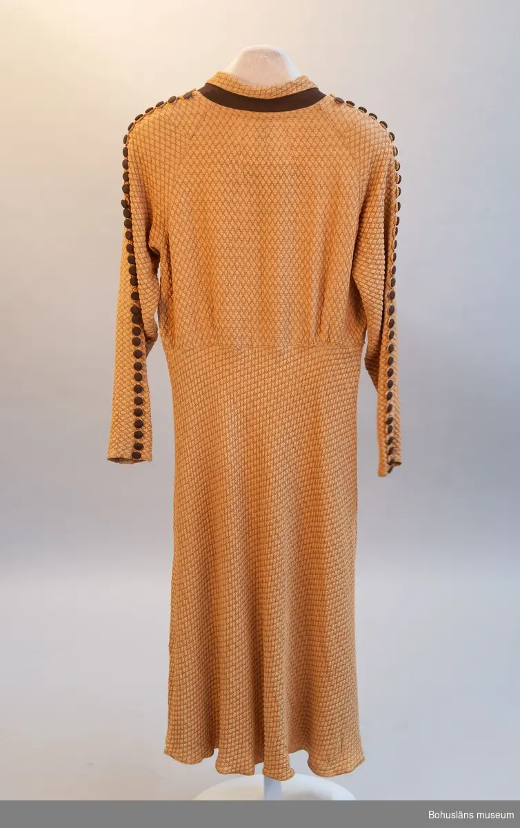Gulbrun klänning med lång ärm och klockad kjol. Krage och klädda knappar längs ärmens överdel.