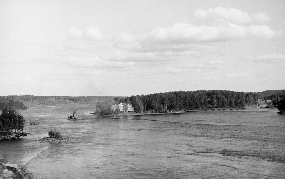 Innsjø ved byen Heinola i  Päijänne-Tavastland (Päijät-Häme) i Finland, fotografert i 1934. Fotografiet er tatt fra et utsiktspunkt - visstnok ei jernbanebu - ned mot en innsjø. Til venstre i bildet ser vi en dampbåt med kurs fra den retningen fotografen sto i. Til høyre for fartøyet, på et nes, ser vi en staselig kvit bygning framfor et skogområdet. Vannspeilet i forgrunnen var lenselagt,, åpenbart fordi dette var en innsjø der det ble buksert tømmer som kunne løsne fra slepene, men ble fanget opp av lensene.