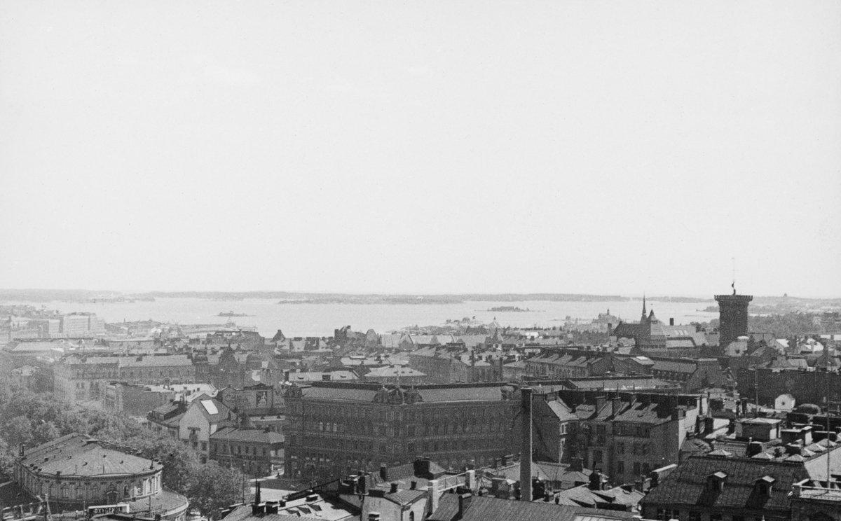 Sentrumsbebyggelse i Helsinki eller Helsingfors, sett fra et høytliggende utsiktspunkt med sjøen og skjærgården i bakgrunnen. Fotografiet ble tatt sommeren 1934, da norske fløtingsfunksjonærer var på studietur i Finland.