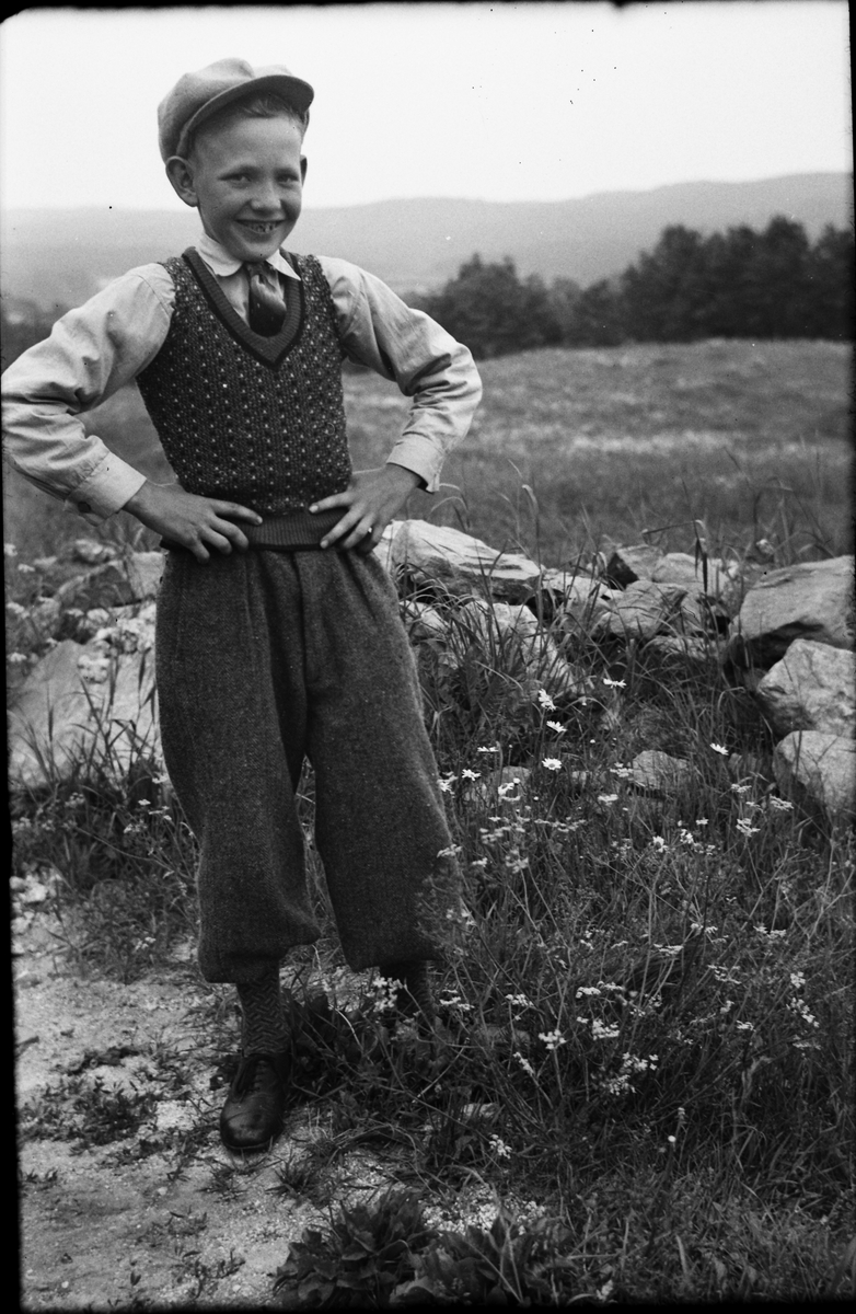 "Husa-Karls" pojk står vid en stengärdsgård klädd i golfbyxor och stickad väst över skjortan med slips och keps.
