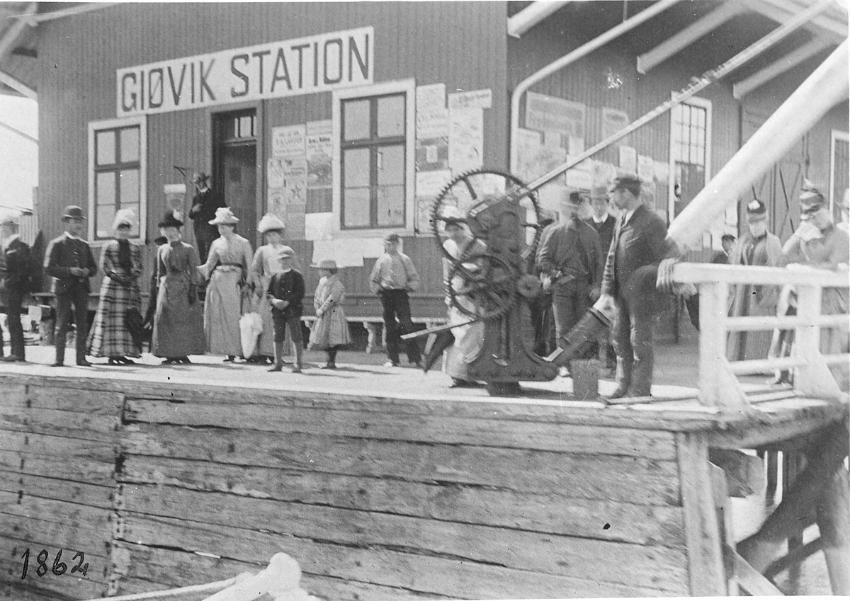 Gjøvik Station som lå på brygga. På brygga sees lastebommen med hådsveiv. Mange personer på brygga som antakelig venter på båt.