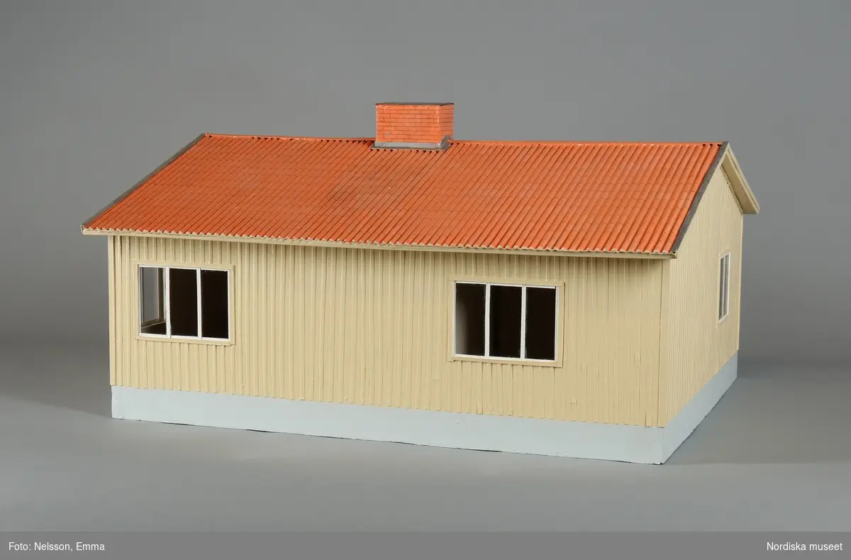 Modell av ett Kölsta-Hus i trä (plywood). Målat. Föreställande a) enplansvilla med gul träpanel och grå grund samt b) avtagbart rött tegeltak. Framtill röd dörr, trapp med räcke, mindre utskjutande tak, två små och ett större fönster, i huset parkettgolv. Två fönster på höger gavel, ett på vänster gavel. Två bredare fönster på baksidan. Skorsten på taket.
/Leif Wallin 2023-08-23