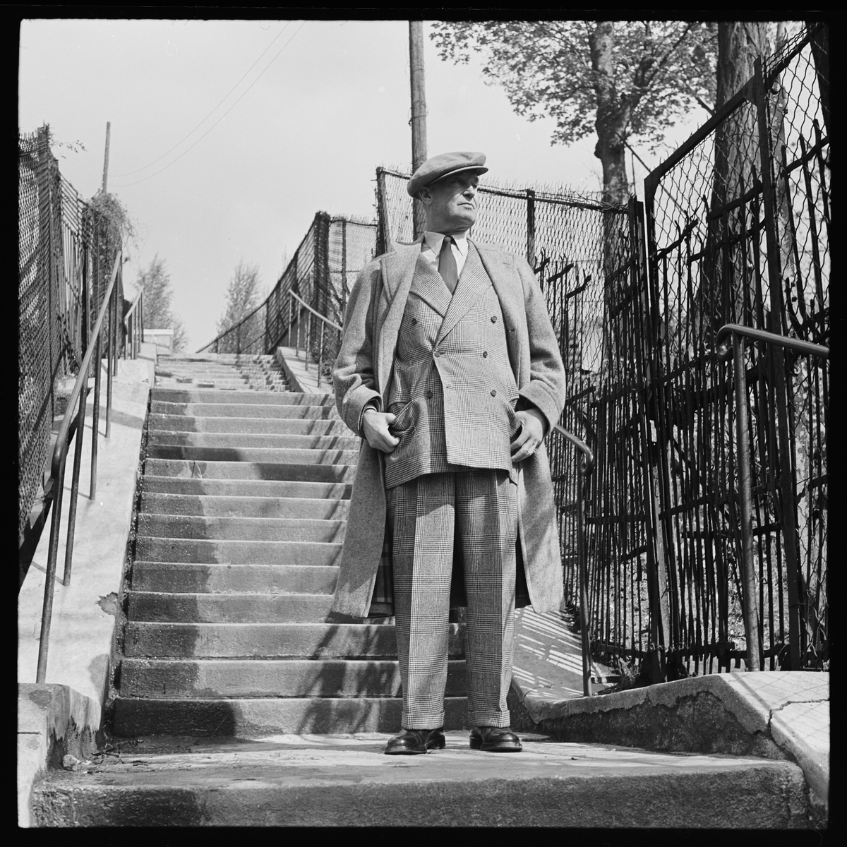 Maurice Chevalier i tweeddress og frakk fotografert i en trapp. 