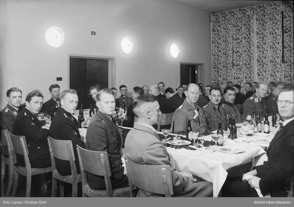 Medlemmer av Germanske SS Norge og tyske soldater og ansatte i det norske politietat? samlet til fest. Øl og dram på bordet (pils fra Sarpsborg bryggeri).
Kan være fra Festiviteten i Sarpsborg (Olavs Hall).
