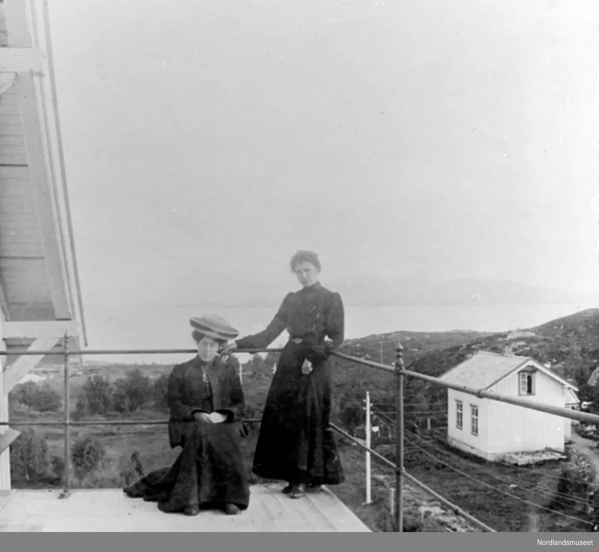 Bildet viser to damer på en altan. Damen til høyre har mørk kjole på seg. Til venstre sittende på en stol er en dame i mørk drakt med stor lys hatt. Til høyre er et hus, ellers stolper og en del busker. Rundt altanen et gelender.