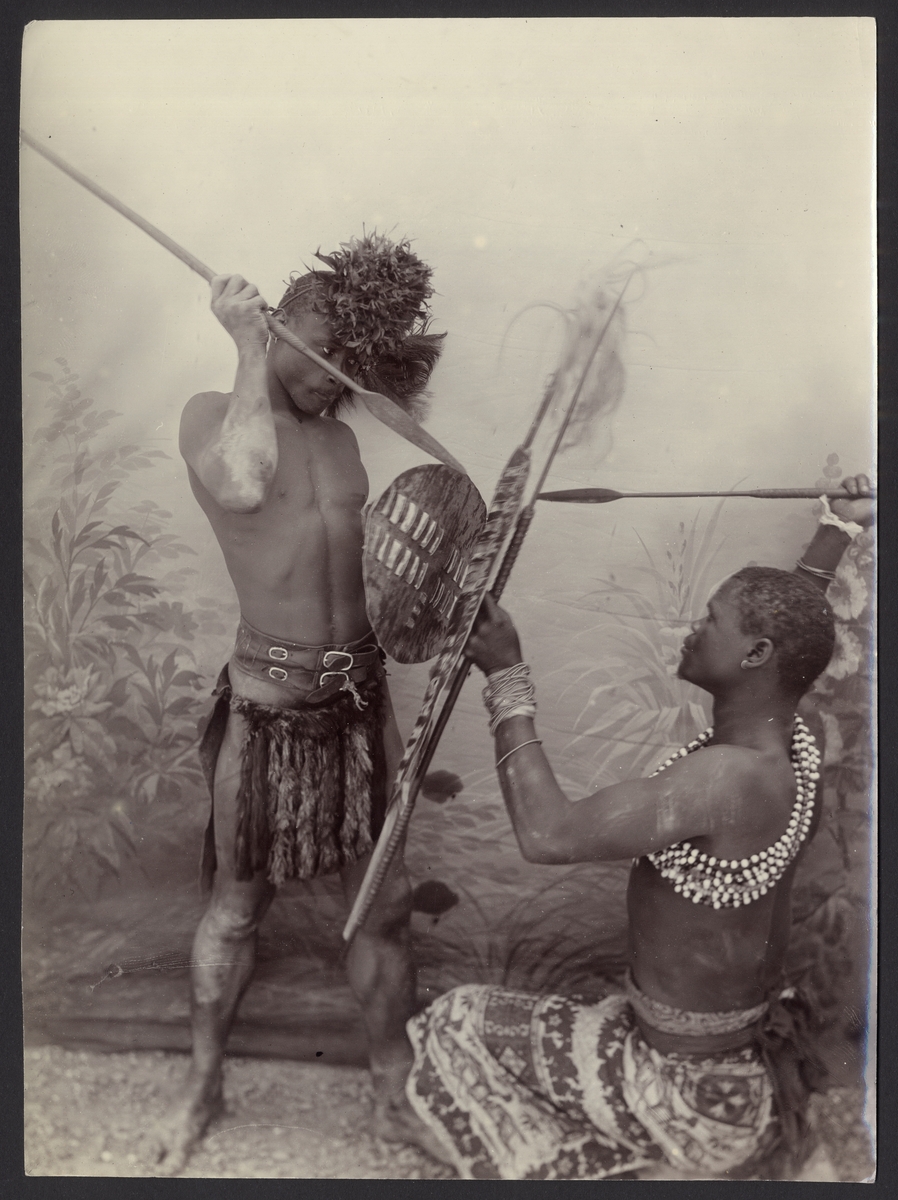 Denna ateljéfotografi föreställer två Zulu-krigare med traditionella kläder och vapen som låtsas strida inför fotokameran.