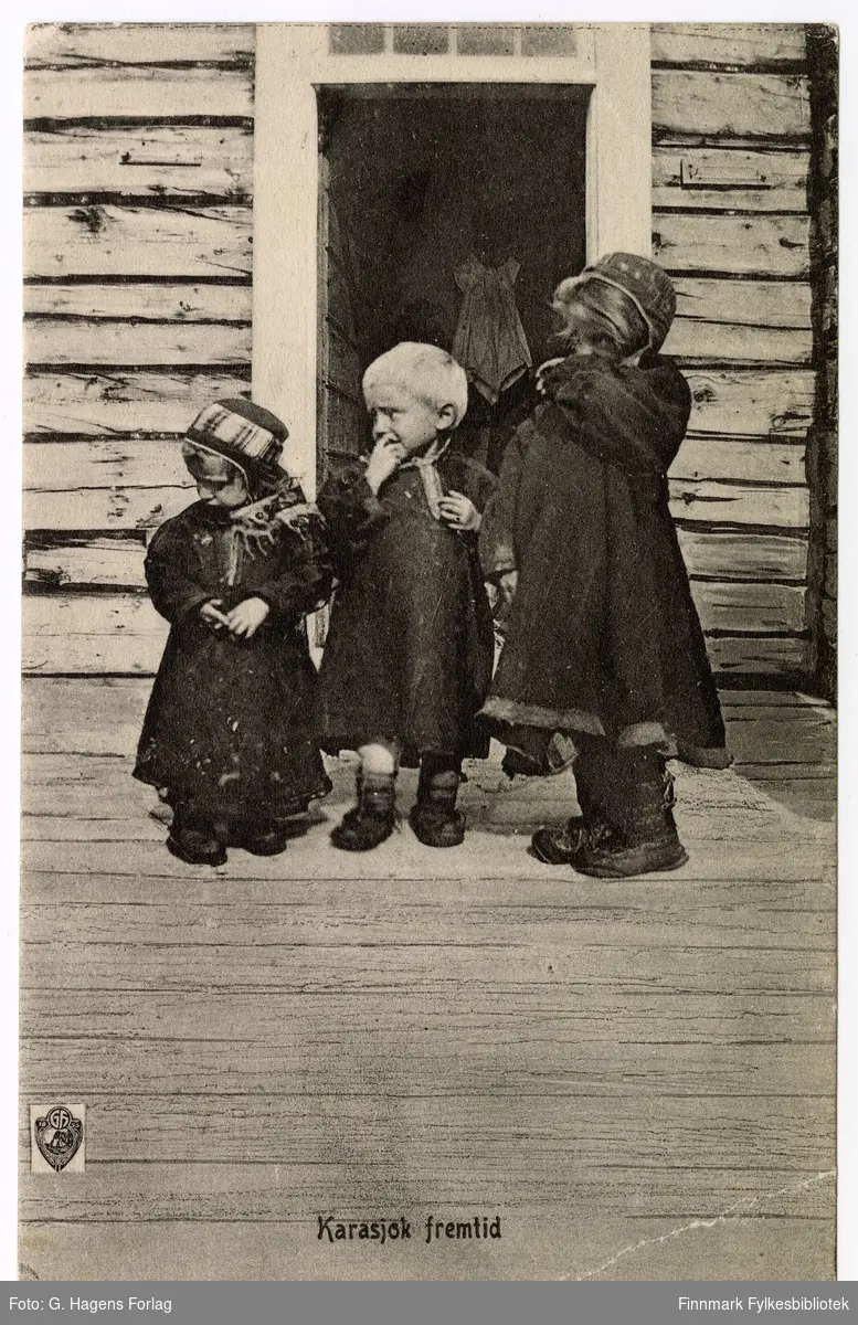 G. Hagen forlag sitt postkort med tittel "Karasjok fremtid". Tre samiske barn står ved døråpningen. En kvinne står inne i huset, bare den lyse forklær er synlig. Dette er en av samemisjonens stasjoner, på bildet ser vi samemisjonenes logo nede til venstre.
