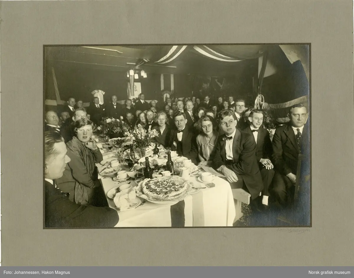I en festpyntet sal sitter mange mennesker til bord dekket med kuverter, blomster og kake.  I bakgrunnen ser vi logoen til Stavanger Lithografisk Anstalt, sammen med tallene 1876-1926.