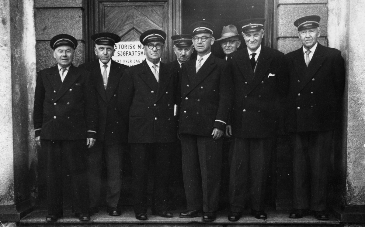 Museumsvakter ved Bergens Sjøfartsmuseum, oppstilt foran døren til kulturhistorisk museum i Bergen. 8 tidligere sjøoffiserer i uniform, samt en sivil med hatt.
