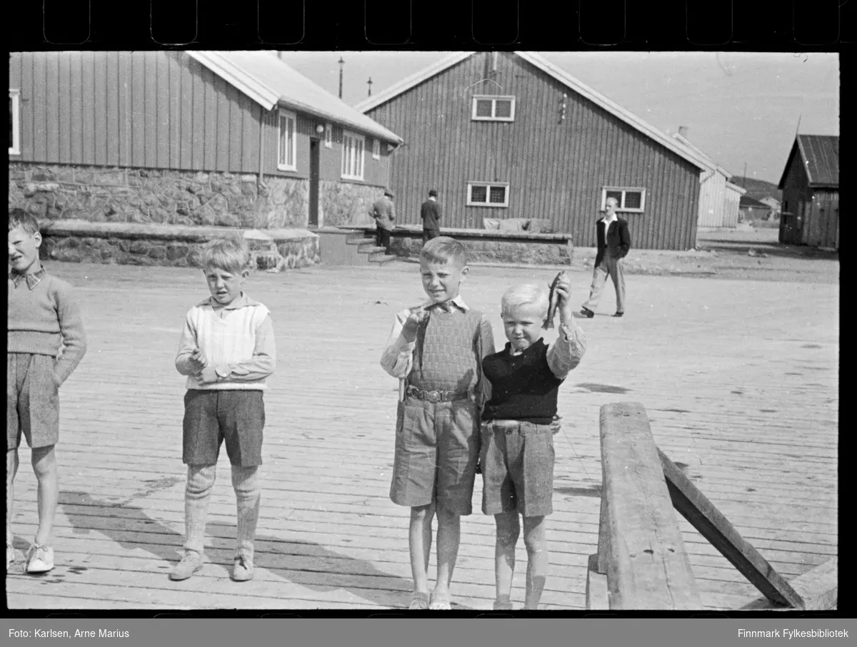 Barn fisker på kai i Kirkenes. En av guttene viser fram en fisk (til høyre)

I bakgrunnen kan man se bygninger

Foto antagelig tatt på slutten av 1940-tallet, tidlig 1950-tallet
