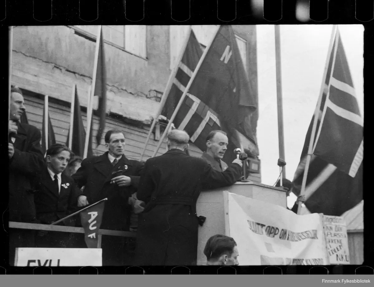Foto av oppmøte ved betongskolen i Kirkenes, antagelig 1. mai 

Foto antagelig tatt på slutten av 1940-tallet, tidlig 1950-tallet 

Man kan se en mann i talerstolen. Ved siden av står en gutt som holder på et AUF flagg