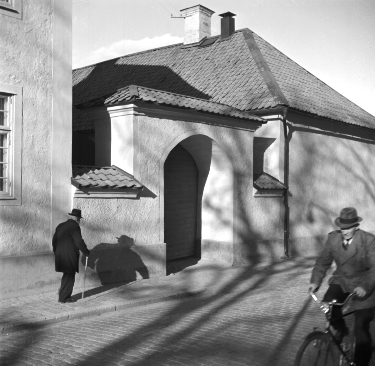 Kvällssolen slog långa skuggor mot Biskopsgårdens fasad när en äldre man snart skulle nå gårdens gatuport. Bilden publicerades i Fotografisk årsbok och hade där titeln "Snart vid porten". Sannolikt finner vi i detta fotografens symboliska tanke. Foto omkring 1950.