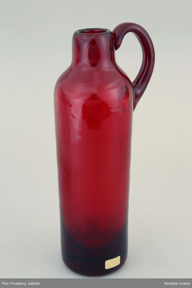 Huvudliggaren:
"Krus av rubinfärgat glas, rak cylindrisk modell, upptill avsmalnande och med öra. Från Reimyre. Design: Monica Bratt."