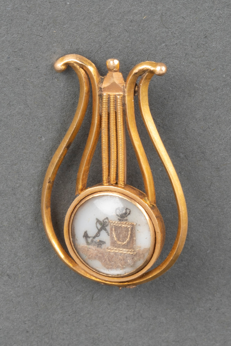 Lyreformet nål i gull. Nederst er det et sirkelrundt felt dekket av glass med dekor på hvit bunn; søylefotstykke med urne og anker.  Nål på baksiden.