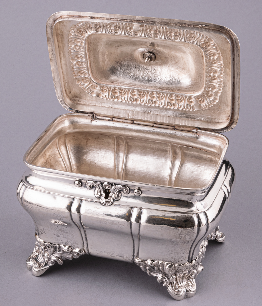 Sockerskrin av silver, fyrsidigt med lock på gångjärn, med nyckel. Uppstående ögleformigt handtag på lockets ovansida.
Stämplad:
E. G. Öström 
G.
V4