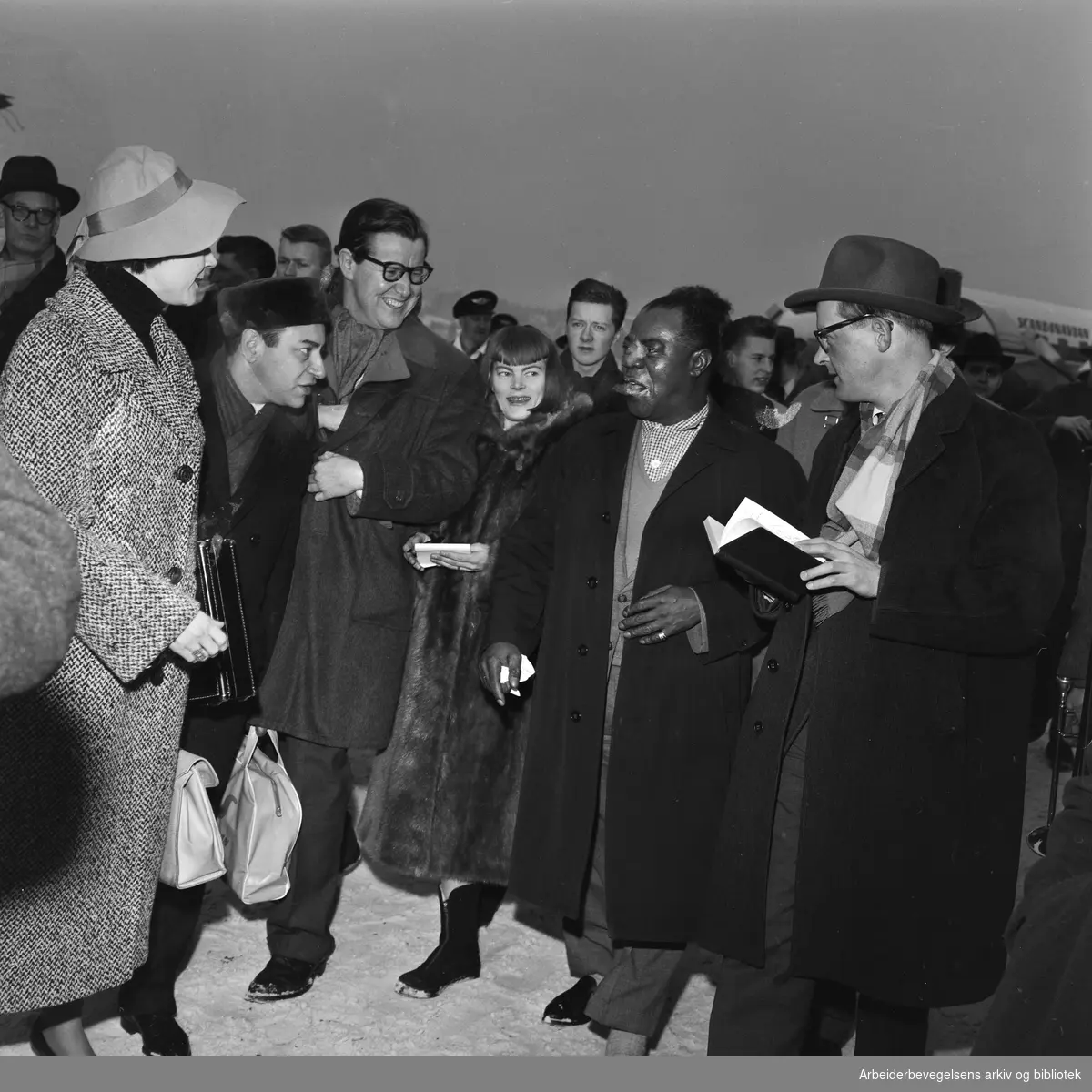 Louis Armstrong ankommer Oslo via Fornebu flyplass 2 februar 1959. Ønskes velkommen og intervuet av blant andre Haagen Ringnes (th) og Tor von Krogh.