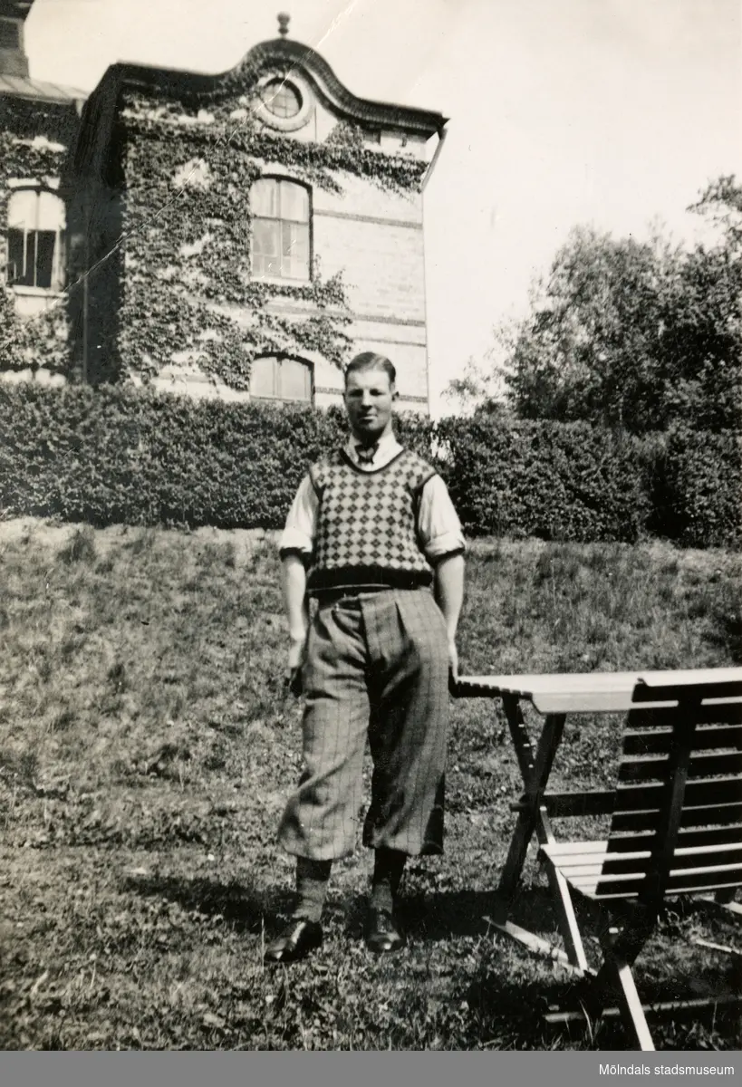 Gustav Berglund står på gräsmattan bredvid en trädgårdsmöbel, 1930-tal. Han arbetade på Stretereds skolhem. I bakgrunden ses Stora skolan där barnen gick i skolan. Byggnaden finns fortfarande kvar (2023).