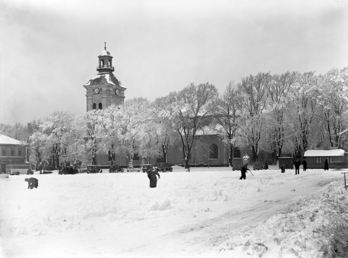 Snöröjning på Varbergs torg med kyrkan i bakgrunden. Framför den står en buss till vänster och en rad parkerade bilar. I snödrivan till höger står en sparkstöttning.