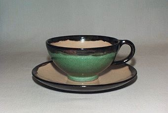 Grön kaffekopp av lergods med gul insida, svart kant och öra. Gult fat av lergods med svart dekorkant och undersida.