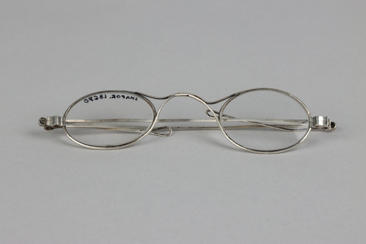 Silver oval eye spectacles, with saddle shaped bridge and lenses. Sliding sides with small loops at the end for a riband.