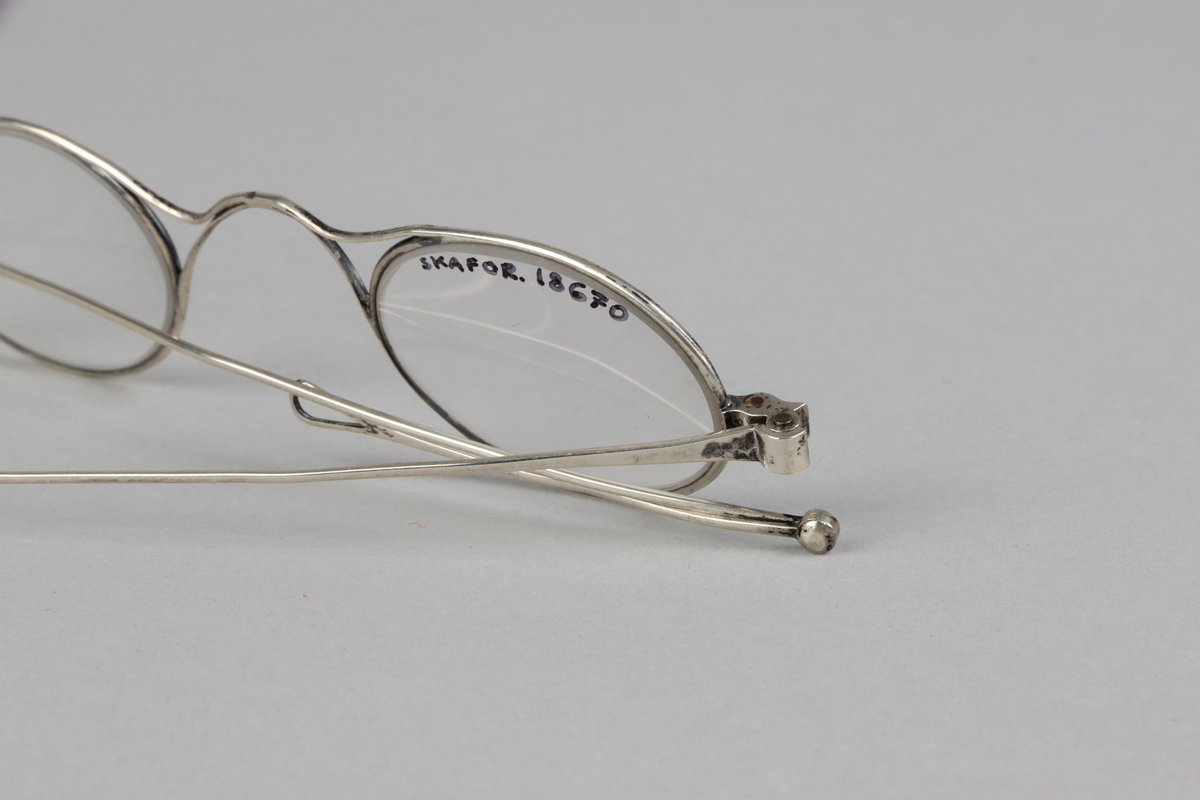 Silver oval eye spectacles, with saddle shaped bridge and lenses. Sliding sides with small loops at the end for a riband.
