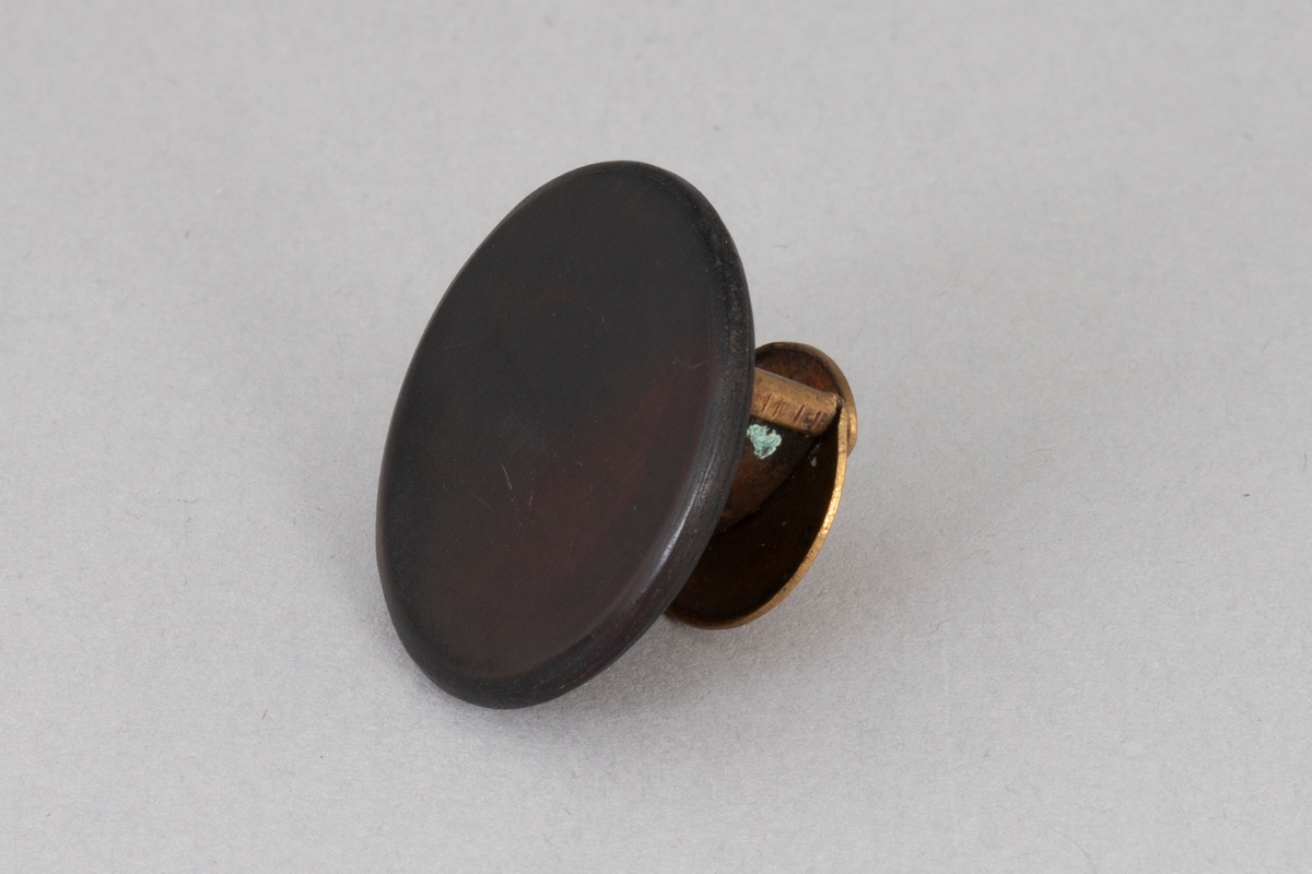 Rund svart knapp i plastlignende materiale med bakside av metall.