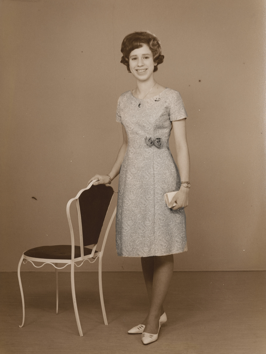 Ung kvinne i kjole i helfigur, stående ved stol.