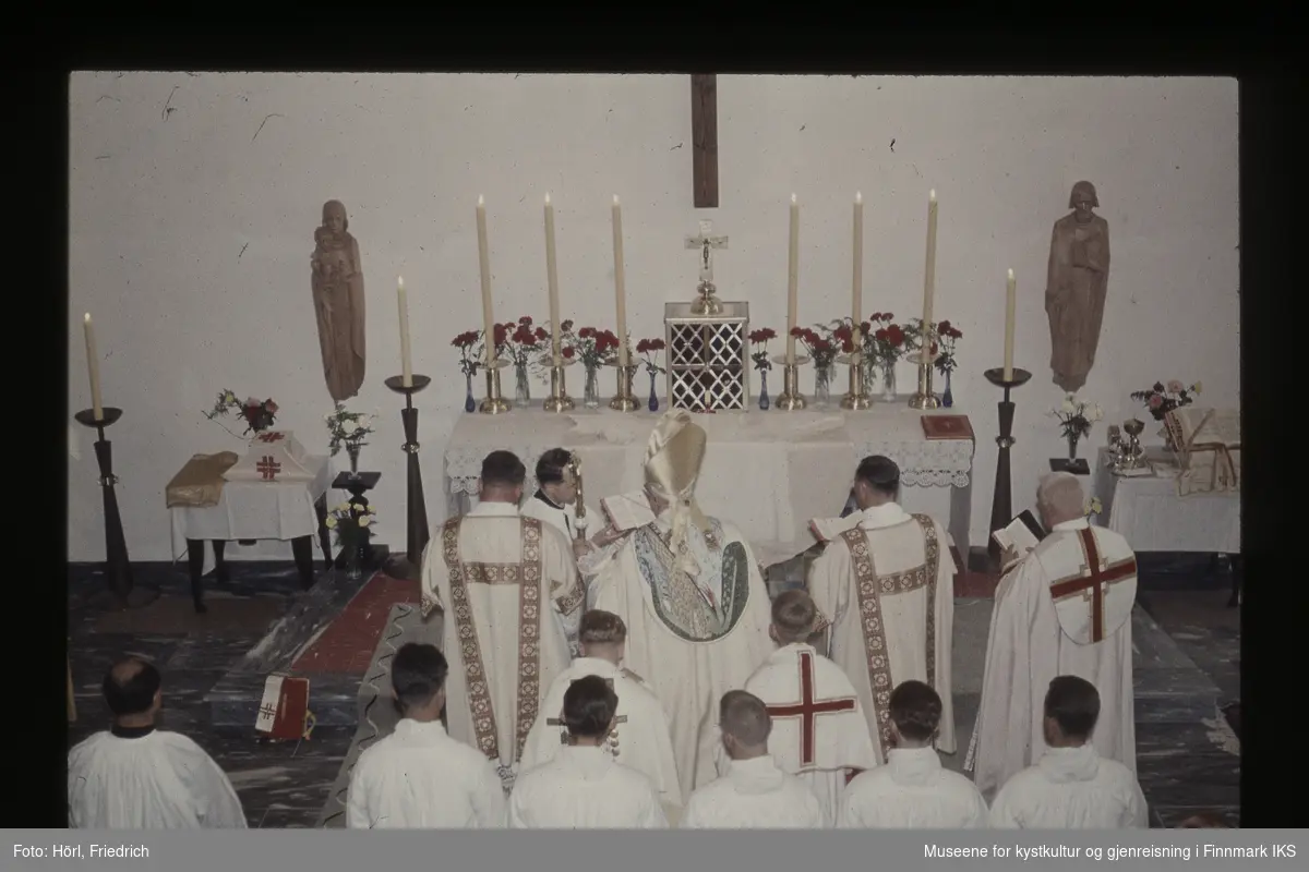 03. august 1958 innvier Biskop Wember Den katolske kirke St. Mikael i Hammerfest. En gruppe seremonielt kledde personer står foran alteret. Ved siden av alteret er det to helgenfigurer av tre på veggen.